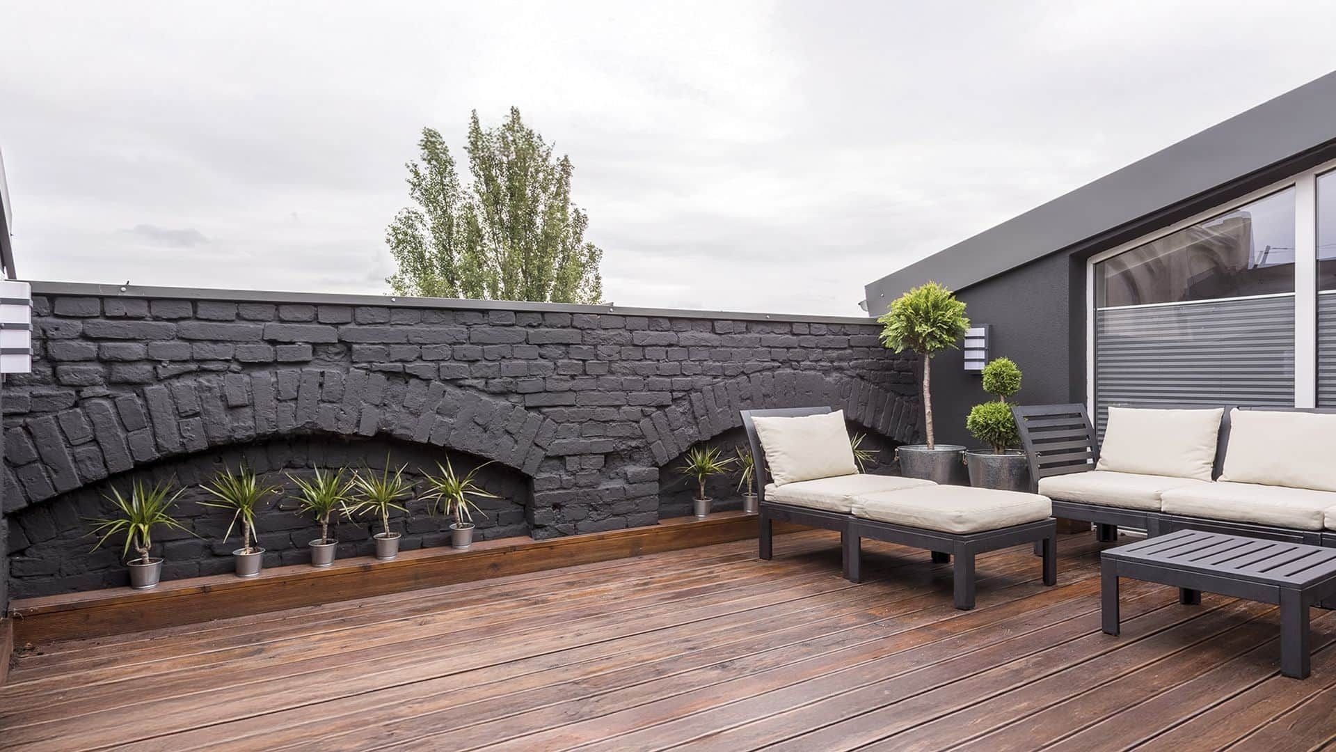 Panoramablick auf eine moderne Dachterrasse mit dunklem Holzboden, Pflanzen, Ziegelzaun und schwarzen Gartenmöbeln